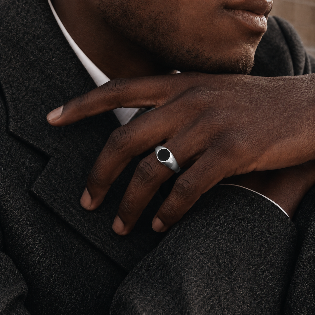 A close-up of a man's hand resting on his lapel, wearing a silver signet ring with a dark oval center stone.