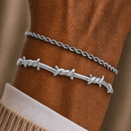 Two silver bracelets worn on a wrist: a thin twisted rope-style chain bracelet and a thicker barbed wire bracelet, shown against a brown skin tone with a beige sweater cuff visible.