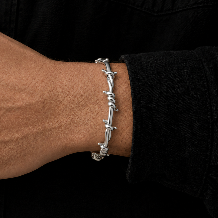 A silver-toned barbed wire bracelet worn on a person's wrist, shown against a black shirt background.