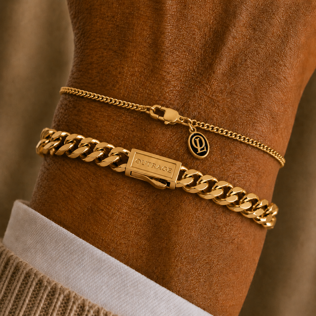 Two gold bracelets, a thick curb chain with a rectangular plate engraved 'O'ORAOE' and a small oval charm, and a thinner chain with a lobster clasp, displayed on a brown mannequin wrist.
