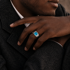 A close-up of a man's hand resting on his lapel, wearing a silver signet ring with a square-cut light blue stone.