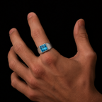 A silver-toned signet ring with a square-cut light blue gemstone, shown worn on a person's index finger against a solid black background.