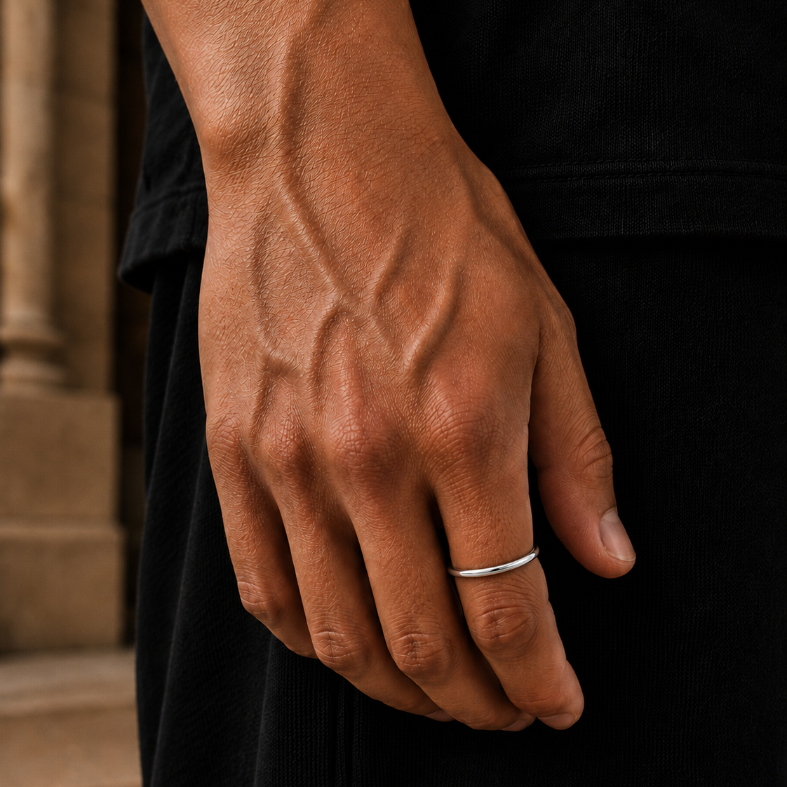 A close-up of a person's hand wearing a thin silver band ring, set against a dark black shirt and a blurred stone background.