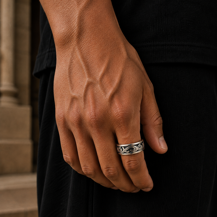 A close-up of a person's hand wearing a silver band ring with a detailed scrollwork design, set against a dark black shirt and background.