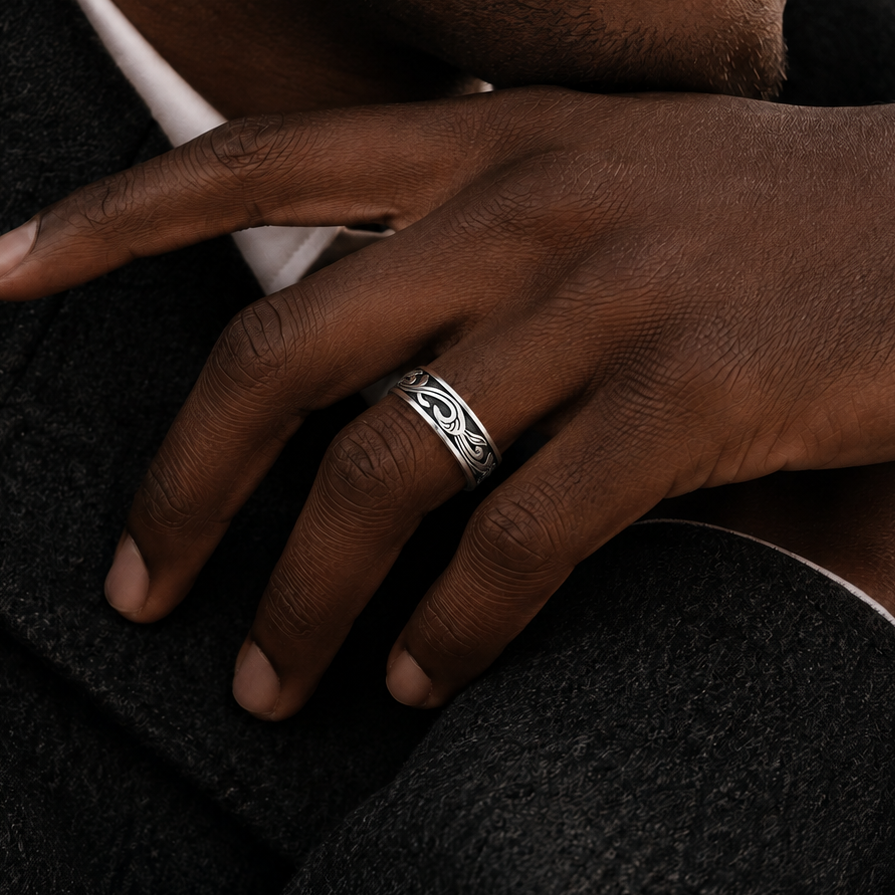 A close-up of a man's hand resting on his lap, wearing a silver band ring with an intricate engraved pattern. He is wearing a dark charcoal gray wool coat over a white collared shirt.