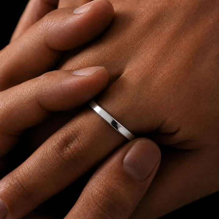A close-up of a person's hand wearing a simple, polished silver band ring on one finger, set against a dark background.