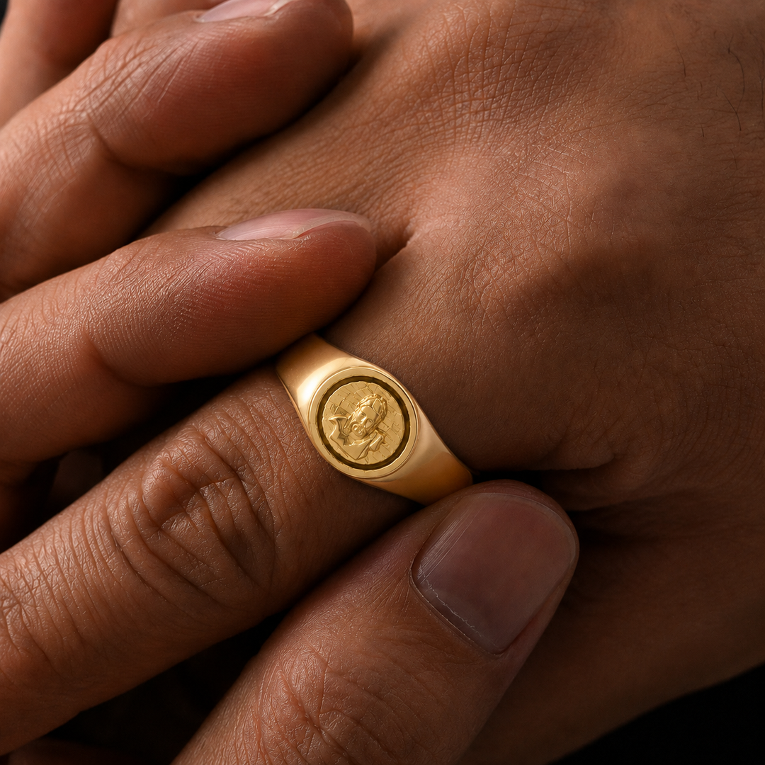 A close-up of a gold signet ring worn on a finger, with the hand resting on another hand against a dark background.