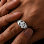 A silver signet ring with an engraved profile bust, worn on a finger and held by another hand against a dark background.