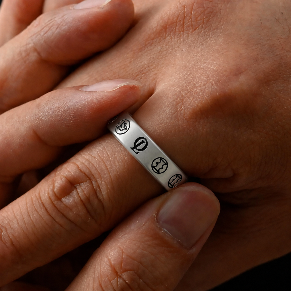 A close-up of a silver band ring worn on a finger, featuring engraved circular symbols, shown against a person's hand.