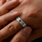 A close-up of a silver band ring worn on a finger, featuring engraved circular symbols, shown against a person's hand.