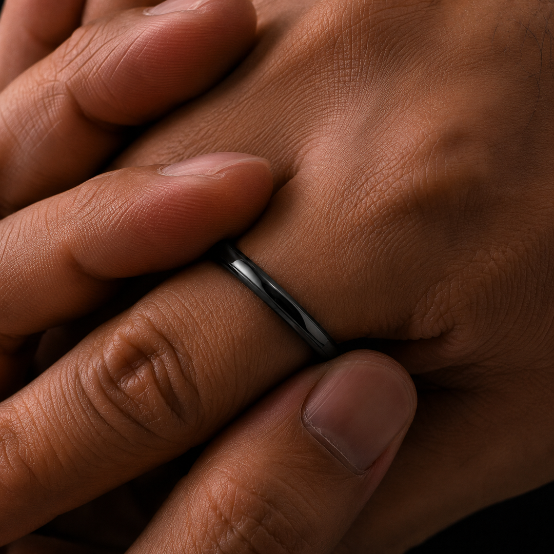 A close-up shot of a sleek, polished black band ring worn on a finger, shown against the backdrop of clasped hands.