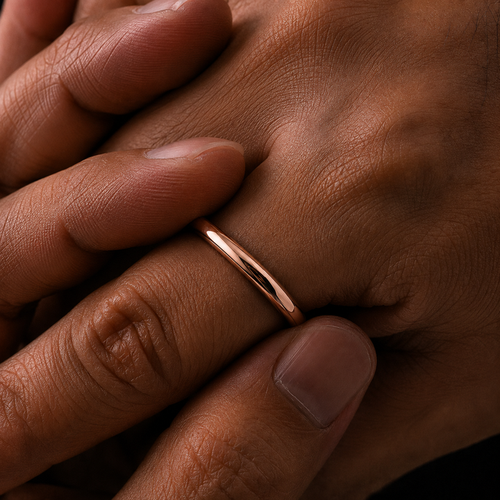 A minimalist rose gold band ring worn on a finger, shown in a close-up lifestyle shot against warm skin tones.