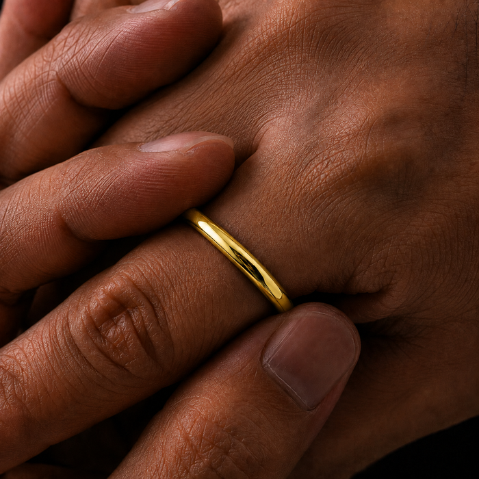 A close-up of a simple polished gold band ring worn on a finger, shown against dark brown skin.