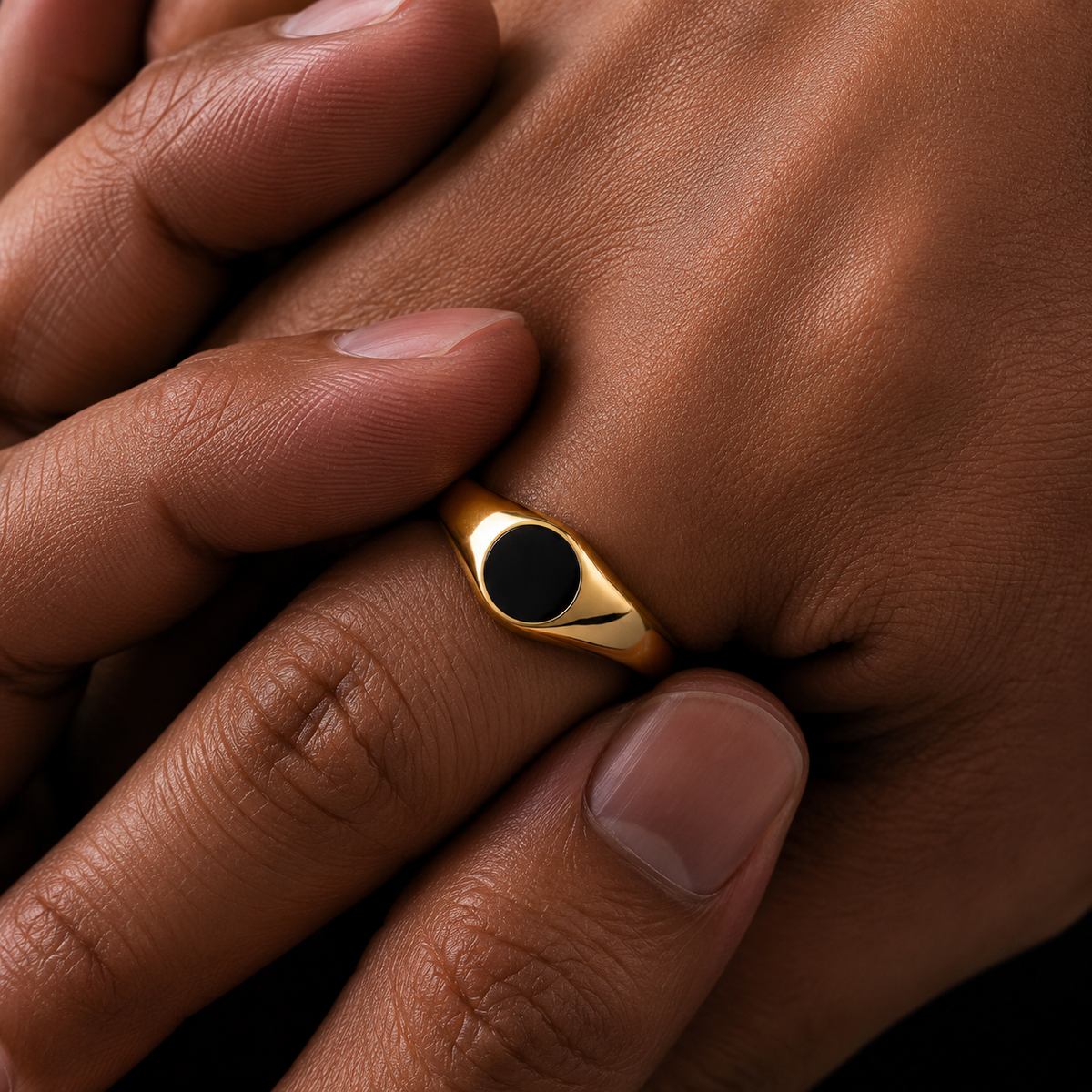 A close-up of a gold signet ring with a black oval face, worn on a person's finger with hands crossed over.