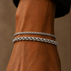 Two silver chain bracelets, one curb link and one twisted rope link, shown on a person's wrist against a warm brown leather background.