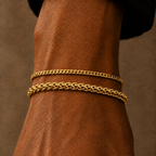 Two gold chain bracelets, one curb link and one curb link, worn stacked on a person's wrist against a warm brown background.