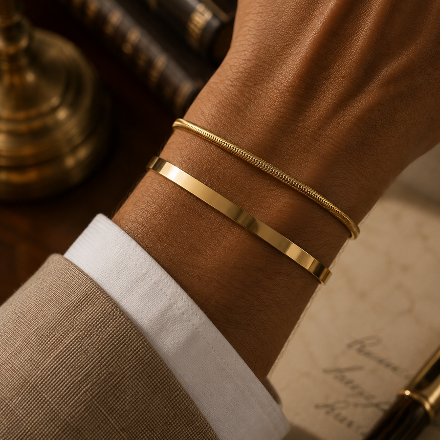 Two slim gold bracelets, one flat band and one textured snake-style bracelet, worn on a person's wrist with a beige suit cuff and white shirt visible.