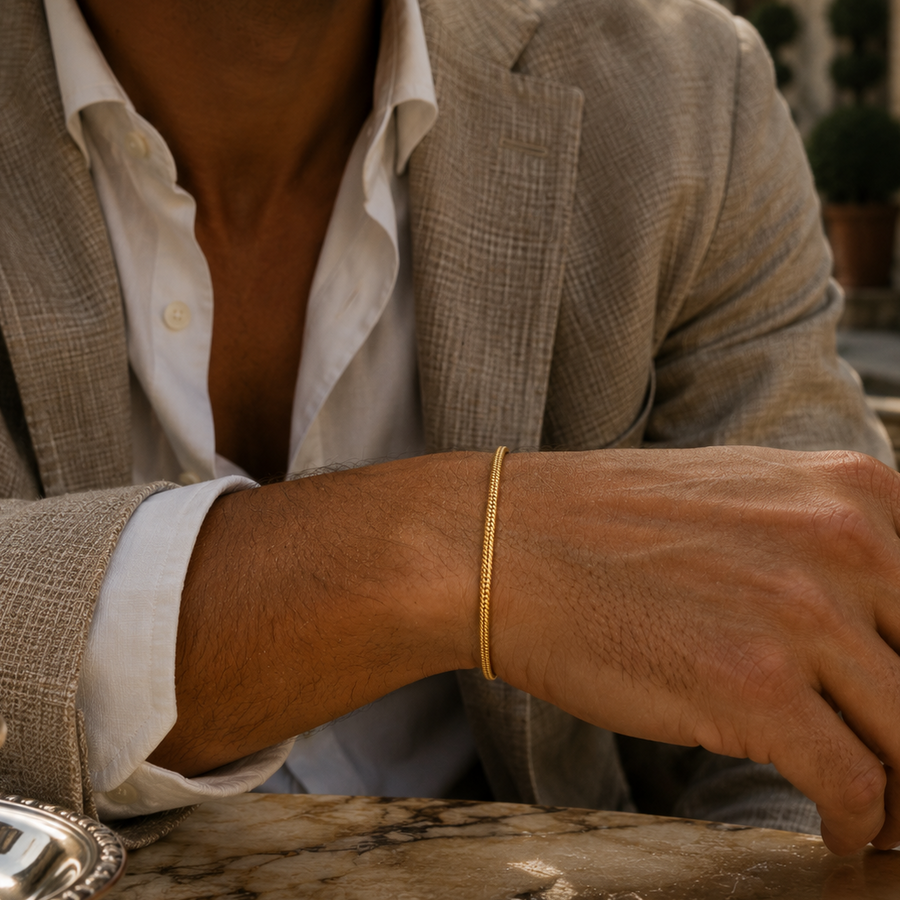 A close-up lifestyle shot of a man wearing a delicate gold bangle bracelet on his wrist, dressed in a light beige blazer and white shirt, seated at a stone table.