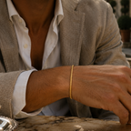 A close-up lifestyle shot of a man wearing a delicate gold bangle bracelet on his wrist, dressed in a light beige blazer and white shirt, seated at a stone table.