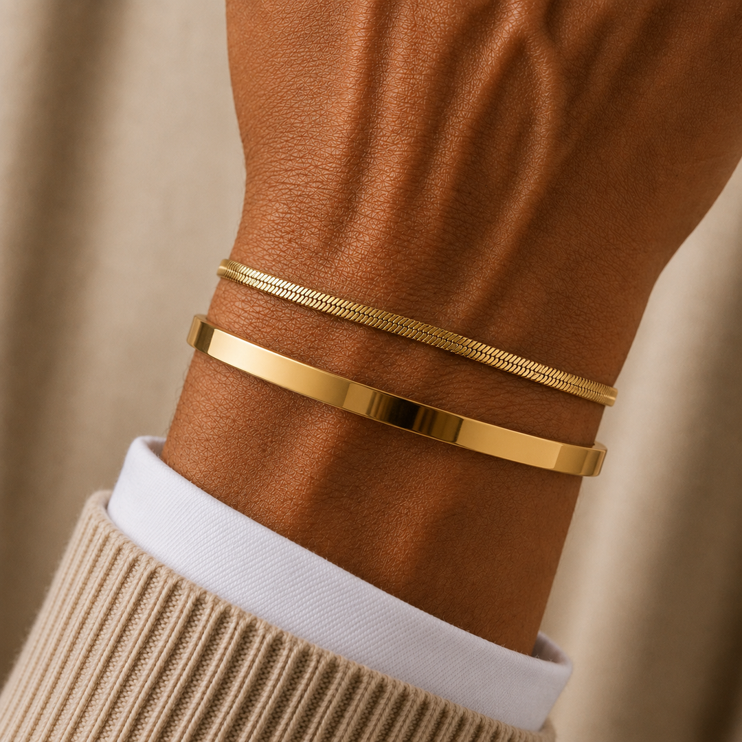 Two slim gold bracelets worn on a person's wrist, with one flat polished band and one textured snake-style band, shown against a warm neutral background.