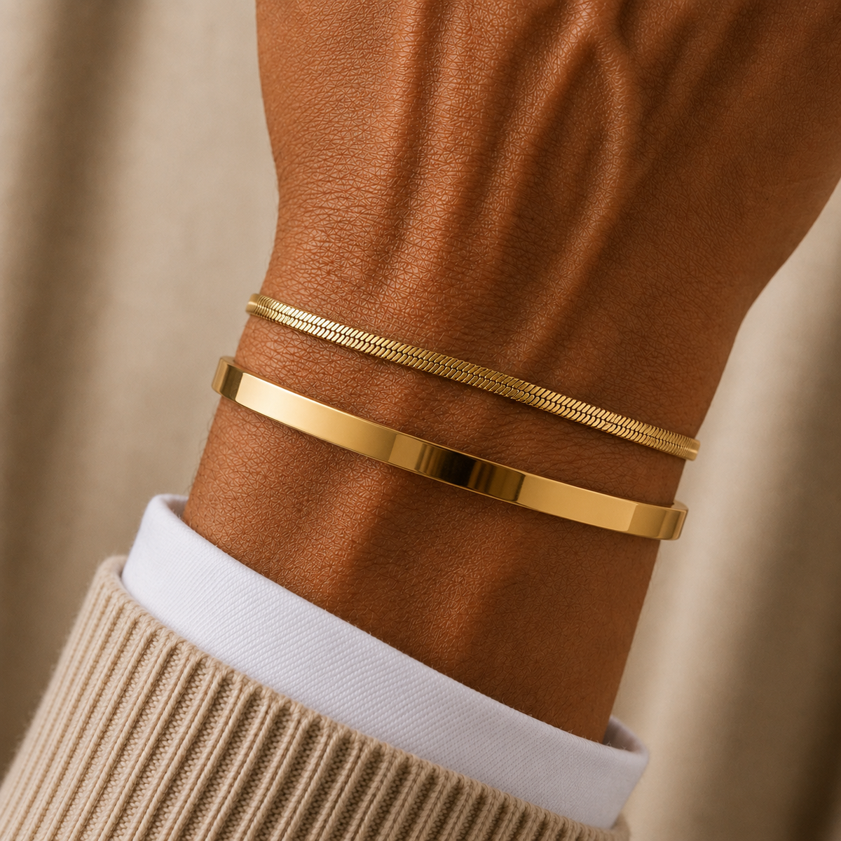 Two slim gold bracelets worn on a person's wrist, with one flat polished band and one textured snake-style band, shown against a warm neutral background.