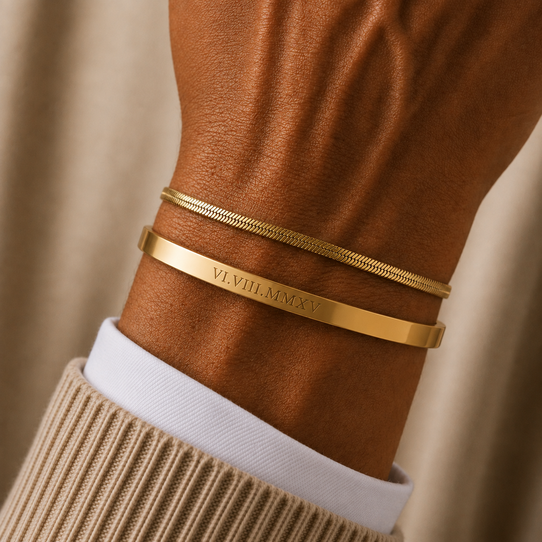 Two gold bracelets worn on a person's wrist, with one flat band engraved with the Roman numerals 'VI VIII MMXV'. The bracelets are styled with a white collared shirt and a beige ribbed sweater against a soft beige background.
