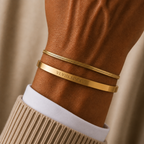 Two gold bracelets worn on a person's wrist, with one flat band engraved with the Roman numerals 'VI VIII MMXV'. The bracelets are styled with a white collared shirt and a beige ribbed sweater against a soft beige background.