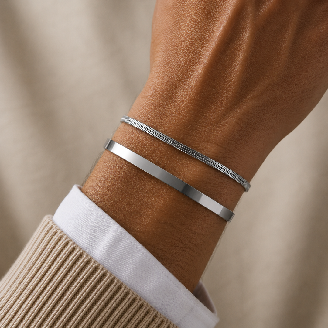 Two silver-toned bracelets, one slim flat band and one textured snake-style bracelet, worn on a person's wrist with a beige sweater cuff and white collared shirt visible.