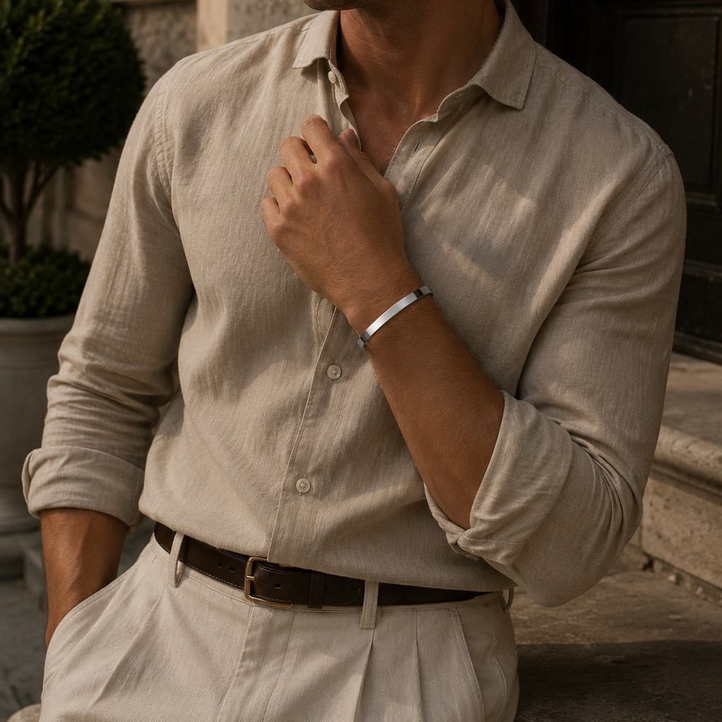 A man wearing a light beige linen button-down shirt and matching trousers, standing outdoors in front of a stone building.