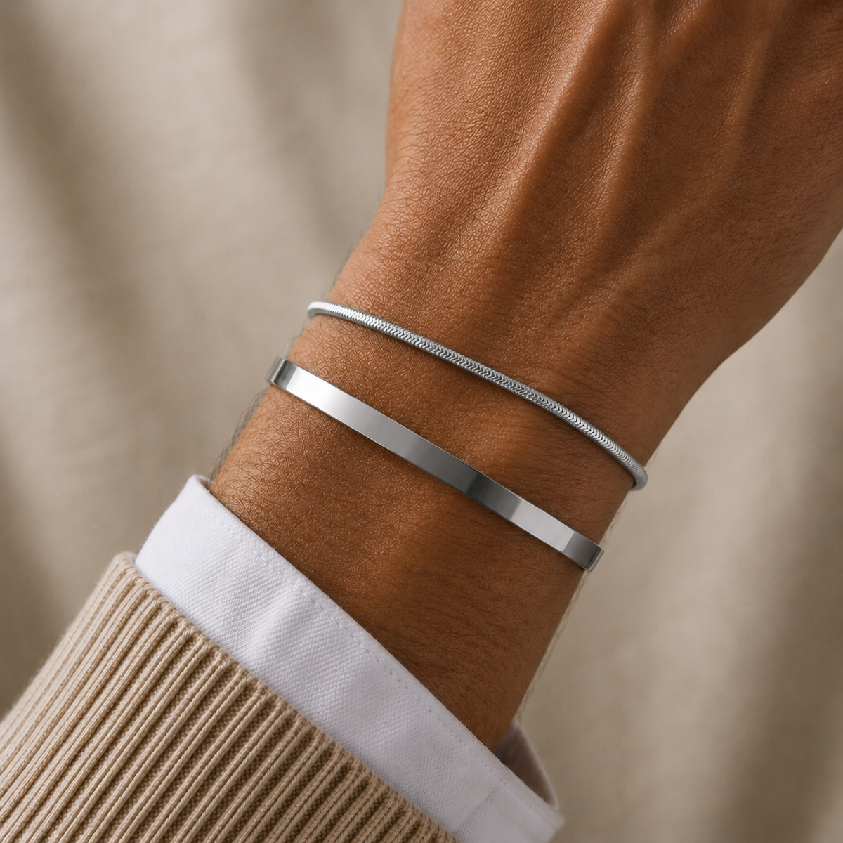 Two slim silver bracelets worn on a person's wrist, with one flat bangle and one snake-style bangle, shown against a soft beige background.