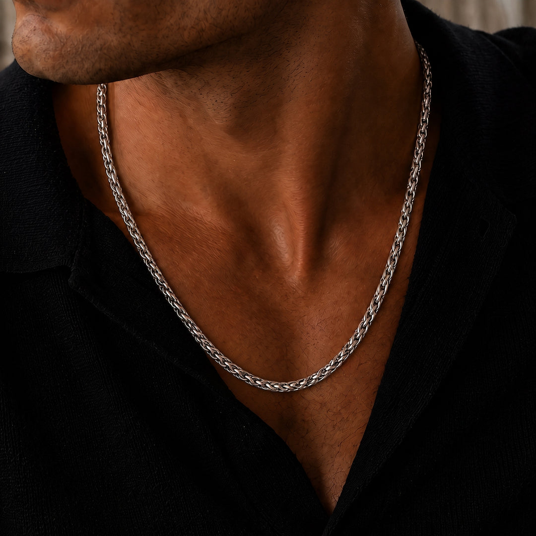 Close-up lifestyle image of a men’s polished silver wheat chain necklace, featuring a tightly woven interlocking link design worn around the neck with an open black collared shirt for a refined, understated luxury look