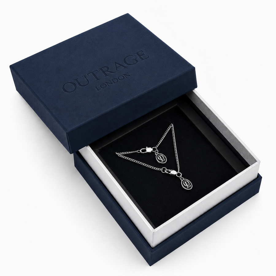 A silver chain necklace with two oval charms, displayed inside an open navy blue Outrage London gift box on a black velvet insert.