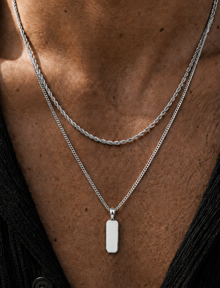 A close-up of a person's neck and chest wearing two layered silver necklaces: one braided chain and one curb chain with a small rectangular pendant.