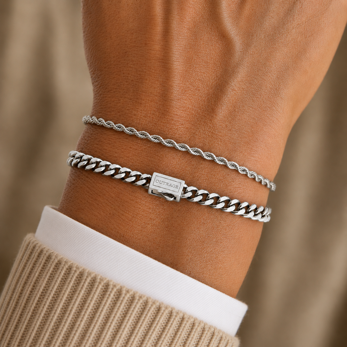 Two silver-toned bracelets on a person's wrist, including a thin twisted rope-style bracelet and a thicker curb-link bracelet with a rectangular brand plaque. The wrist is shown wearing a beige knit sweater over a white shirt cuff, against a soft beige background.