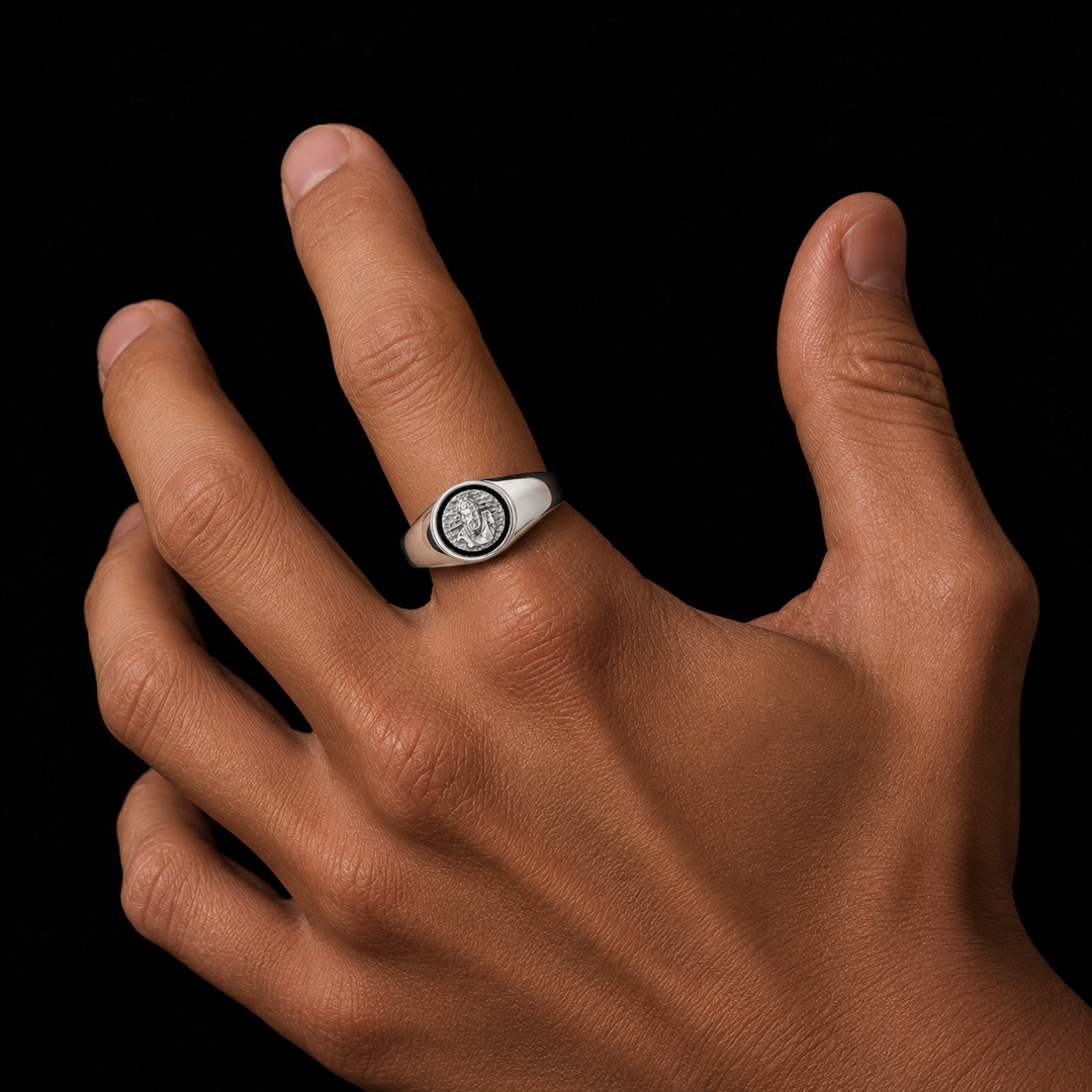 A silver signet ring featuring a circular black-and-white emblem, worn on the ring finger of a hand against a solid black background.