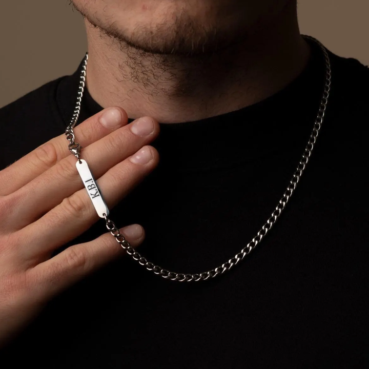 A close-up of a man wearing a silver chain necklace with a rectangular engraved tag that reads 'JEF'.
