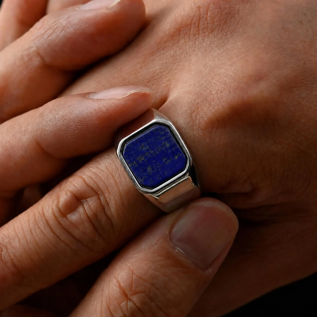 A silver signet ring with a square blue lapis lazuli stone featuring gold astrological symbols, shown worn on a finger.