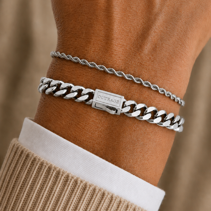 A close-up of a person's wrist wearing two silver bracelets: a thin twisted chain and a wider curb-link style bracelet with a small rectangular brand plate. The person is wearing a beige ribbed sweater over a white collared shirt, against a soft beige fabric background.