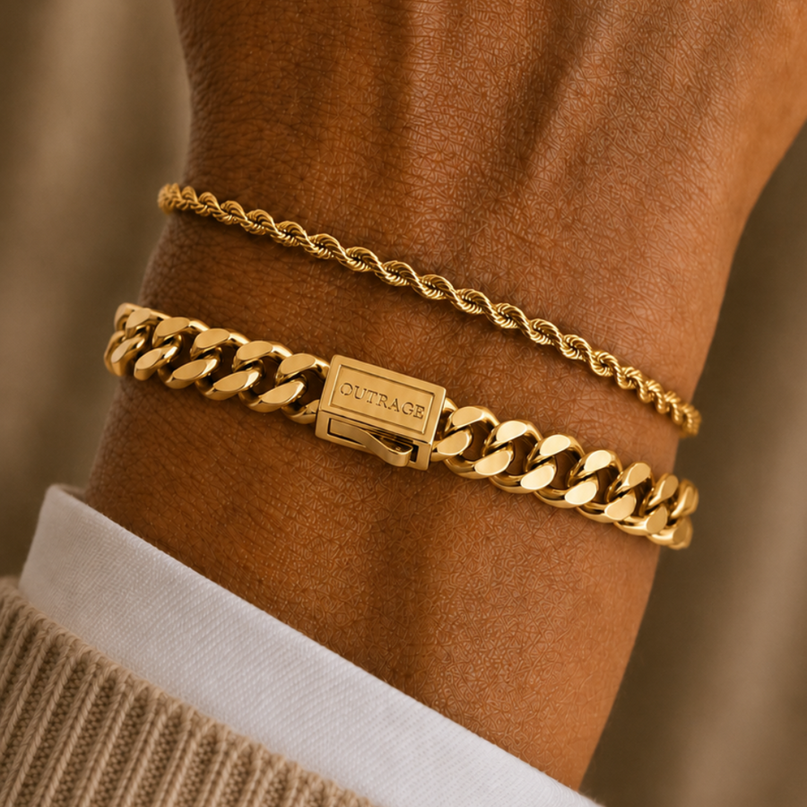 A close-up of a person's wrist wearing two gold bracelets: a delicate cable chain and a thicker curb link bracelet with a rectangular clasp engraved with 'TROY'.