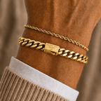 A close-up of a person's wrist wearing two gold bracelets: a delicate cable chain bracelet and a thicker curb link bracelet with a clasp engraved with the brand name 'JEWELRY CRAFT'.