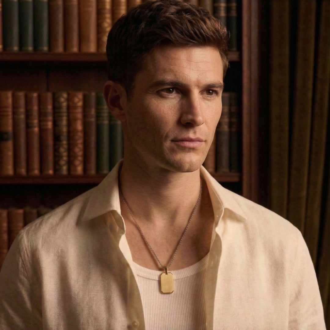 A man wearing a white shirt and a necklace stands in front of a bookshelf.