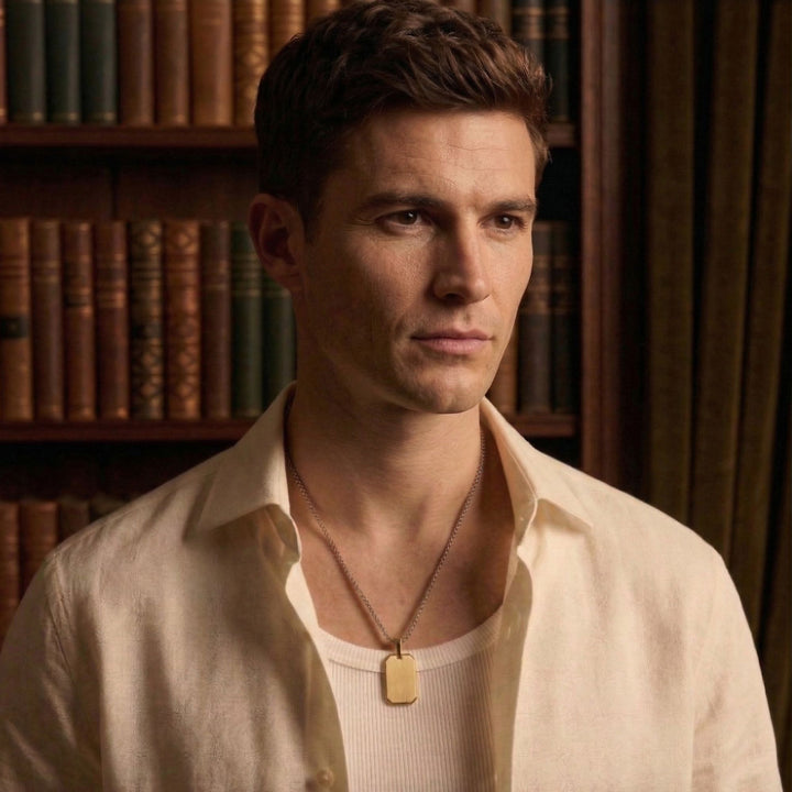 A man wearing a white shirt and a necklace stands in front of a bookshelf.