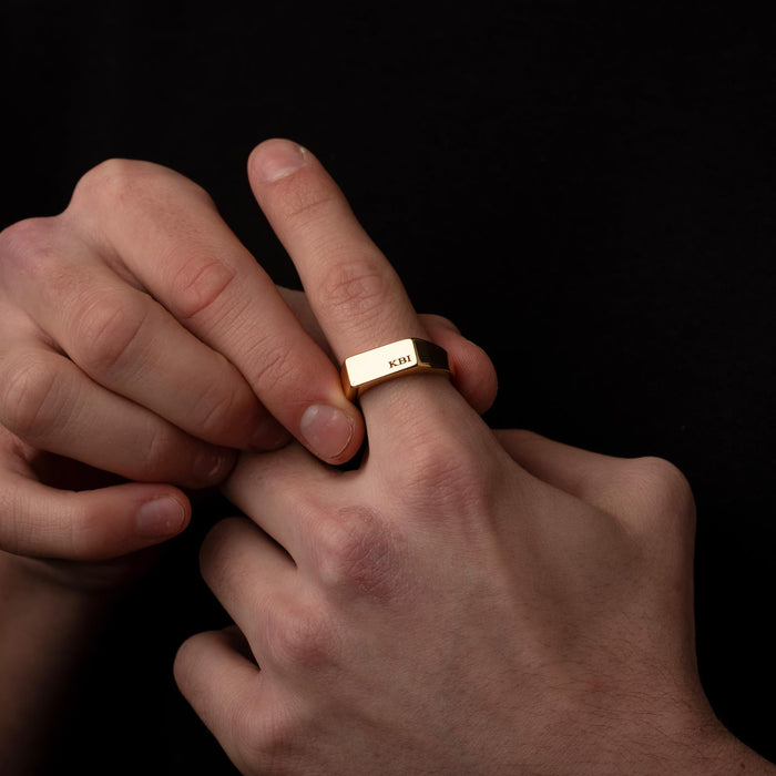 Close-up of a hand wearing a gold ring with 'KBI' engraving on a black background