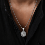 A silver pendant necklace featuring an engraved circular medallion, worn on a model's neck over a black collared shirt.
