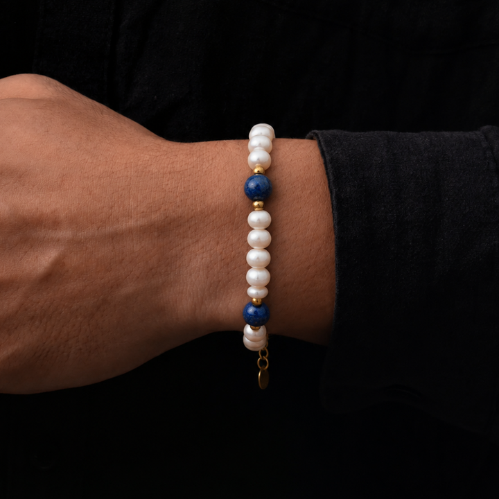 A close-up of a person's wrist wearing a beaded bracelet with white pearl-like beads, two dark blue beads, and small gold spacer accents, set against a dark black shirt background.