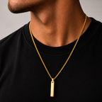 A close-up of a man's neck and chest wearing a gold chain necklace with a slim rectangular gold pendant, paired with a black crewneck t-shirt against a neutral gray background.