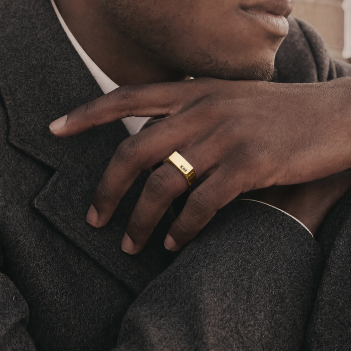 A close-up lifestyle shot of a man wearing a gold signet ring engraved with 'JESUS' on his ring finger, dressed in a dark gray wool blazer over a white collared shirt.