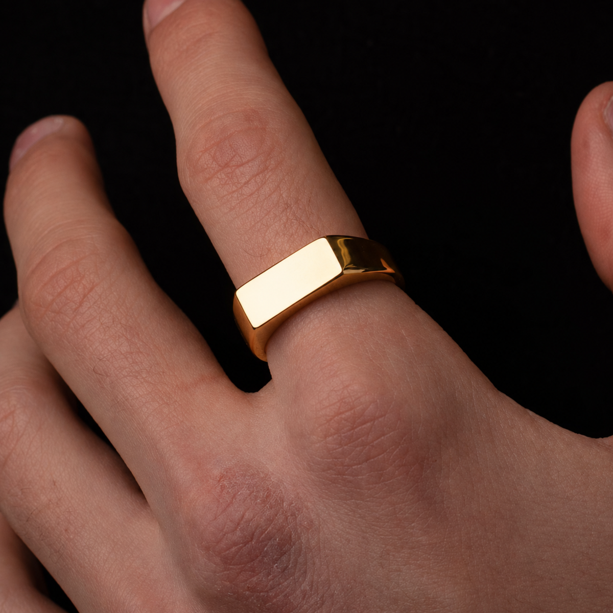 A polished yellow gold signet ring with a flat rectangular face, shown worn on a person's finger against a solid black background.