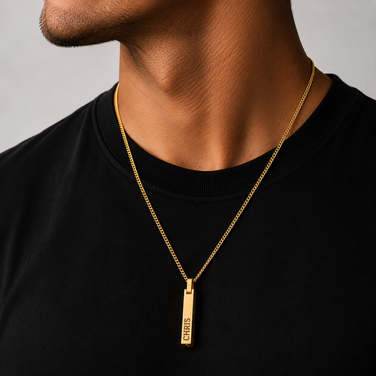 A gold bar pendant necklace engraved with 'CHRIS' is worn by a man in a black crewneck shirt against a neutral gray background.