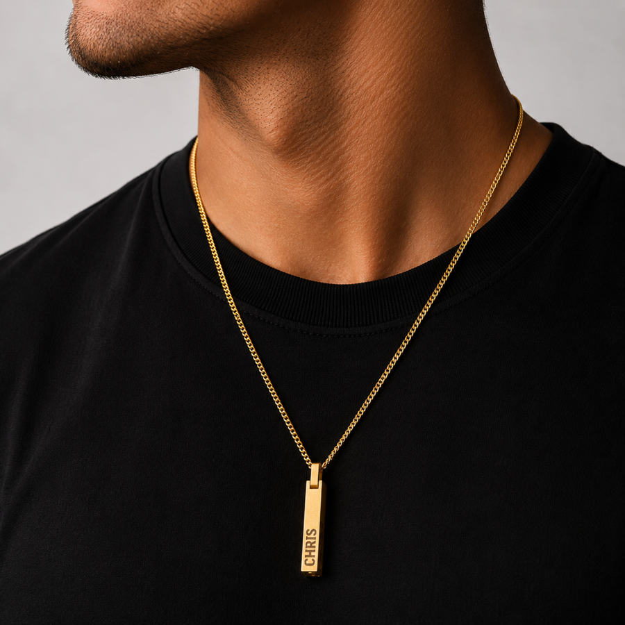 A gold bar pendant necklace engraved with 'CHRIS' is worn by a man in a black crewneck shirt against a neutral gray background.